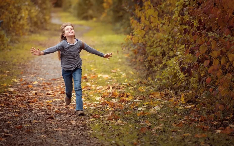 Young girl with long blond hair running along a forest trail smiling as she looks up at the sky.