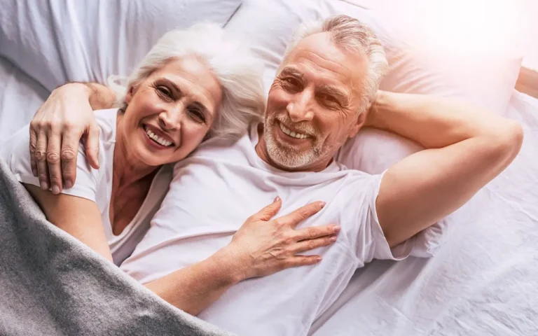 Elderly couple lying in bed together in the morning, smiling beautifully at the camera.