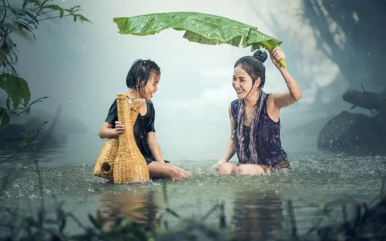 Asian mother and young daughter sitting in tropical river together. Mother is hold banana leaf above them to act as umbrella from the rain.