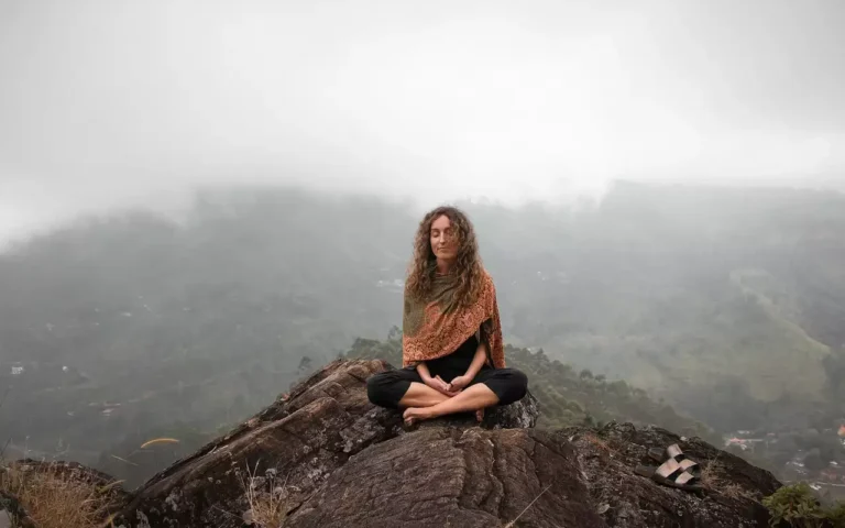 Woman in meditation posture on rocky mountain top as soft morning clouds float through the elevated landscape at dawn